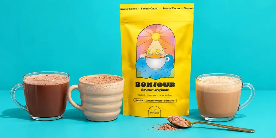 Busta gialla di Bonjour Drink tra tre tazze di bevanda al cacao di tonalità diverse e un cucchiaino di polvere, su sfondo turchese
