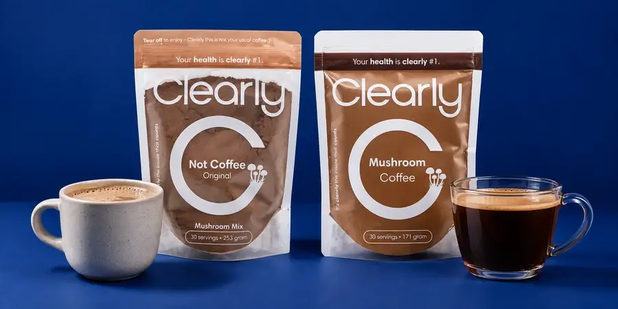 Le buste di Clearly Not Coffee e di Clearly Mushroom Coffee tra una tazza di bevanda cremosa e una tazza di caffè nero, su sfondo blu notte